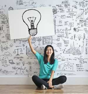 Asian woman sits in front of whiteboard covered by illustrations holding up a large lightbulb drawn on a white piece of paper
